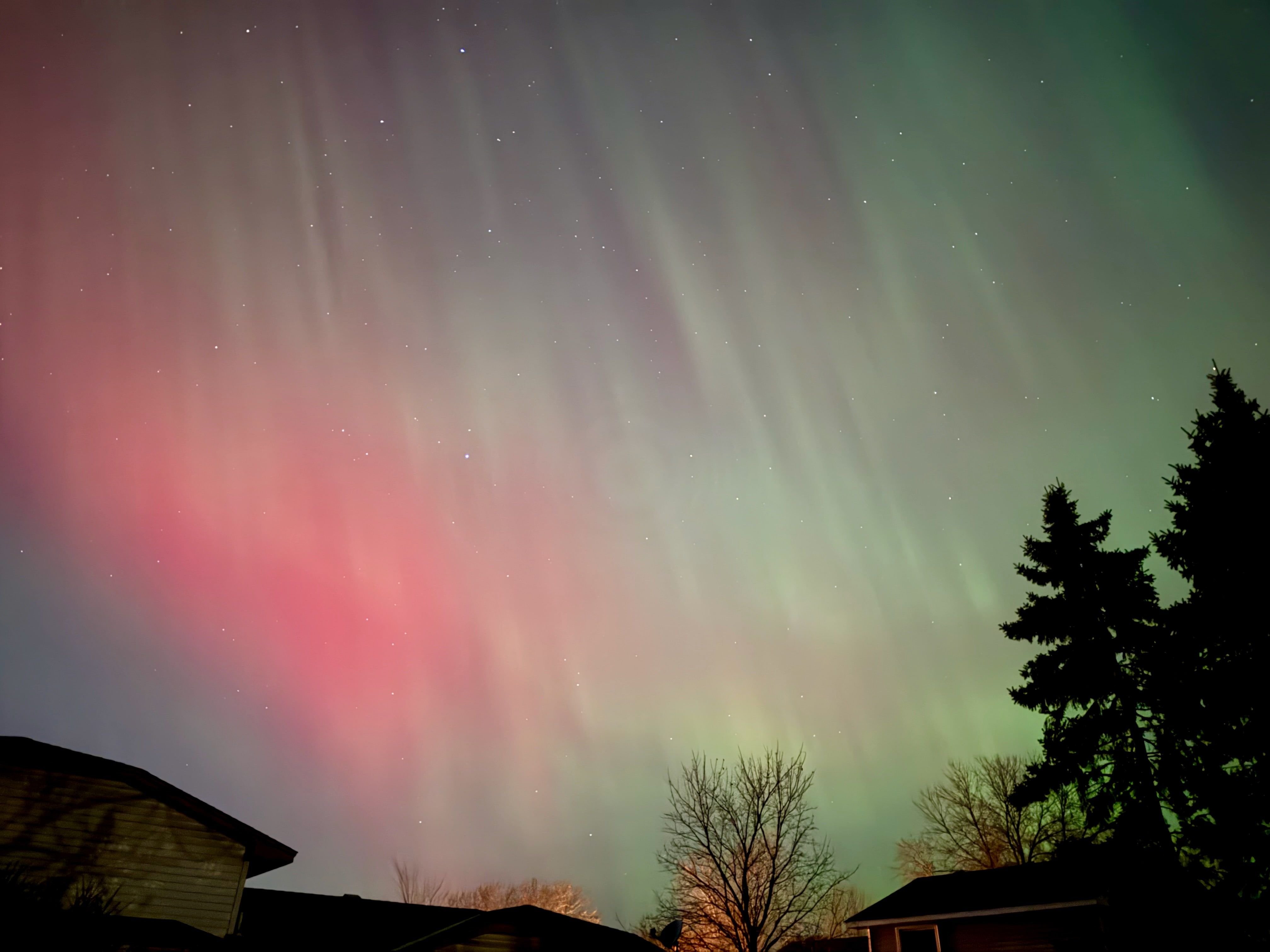 Red and green aurora filling the night sky above a rooftop and bare winter treeline