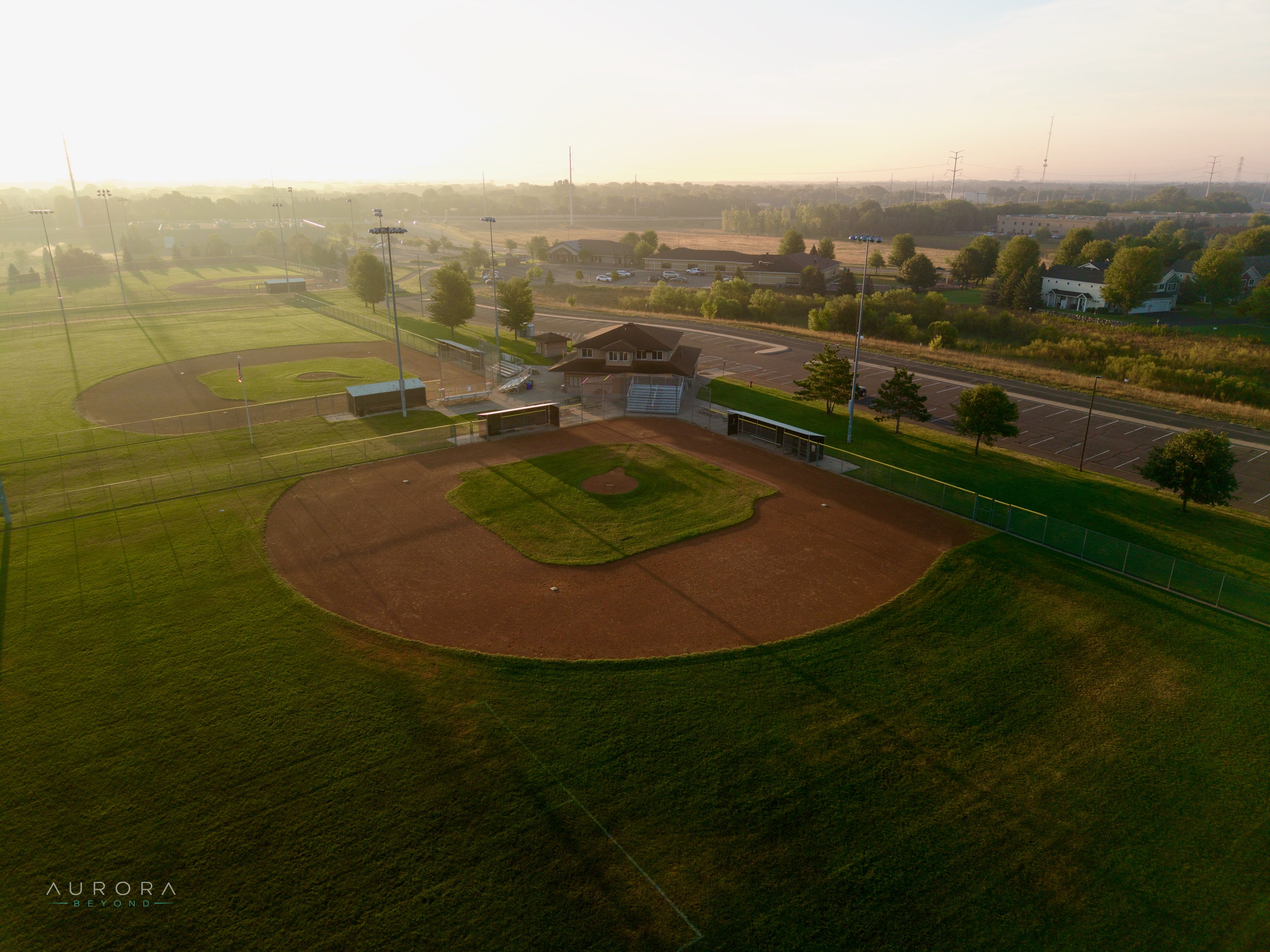Aerial view of an empty ballfield at sunrise, its infield in long shadow, with a second diamond, a park building, and hazy suburban treeline beyond.