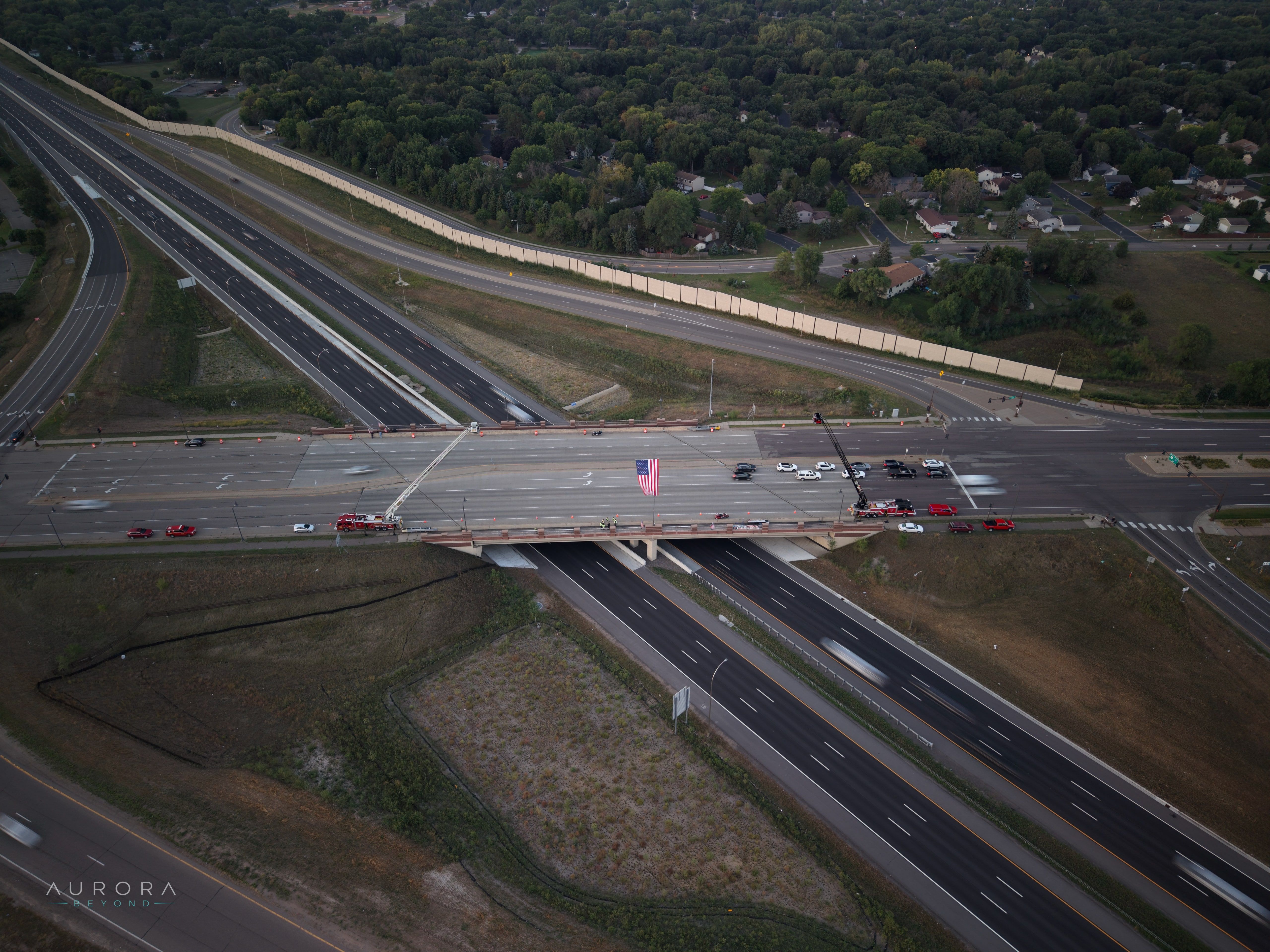 Aerial view of a large American flag hung between two extended fire truck ladders over a highway overpass at dusk, with traffic passing below.